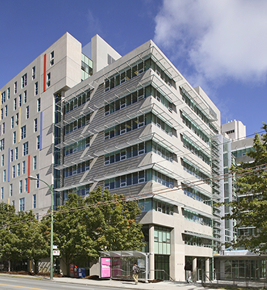 Modern multi-story building with angular windows and trees in front, under a blue sky with some clouds.