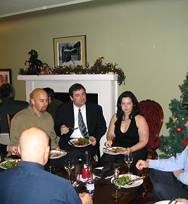 Five people sit around a table eating dinner, with a decorated Christmas tree and mantel in the background.