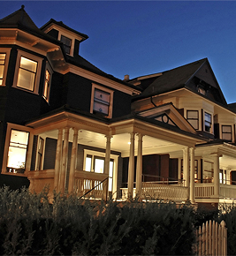 Large, two-story Victorian house with a wraparound porch, lit warmly at dusk against a dark blue sky.