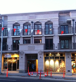 A modern, three-story building with shops on the ground floor and balconies above, orange cones on sidewalk.