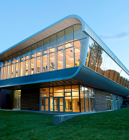 Modern, two-story glass building with curved edges, glowing lights inside, and green lawn in front.