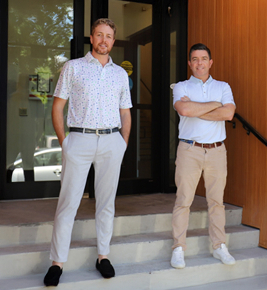 Two men stand on steps outside a building, both wearing light-colored pants and short-sleeve shirts.