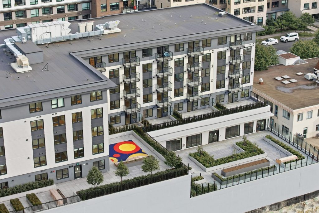 Aerial view of a modern residential building with a landscaped rooftop courtyard featuring raised garden beds, trees, seating areas, and a colourful children's play area