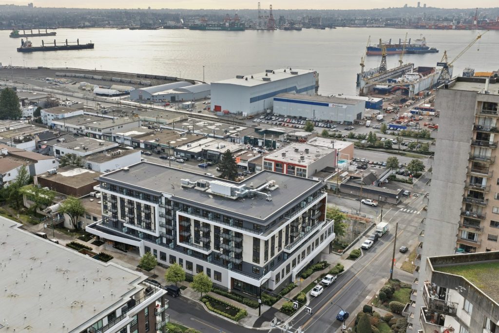 Aerial view of a modern mid-rise residential building in North Vancouver surrounded by the city streetscape, industrial port, and Burrard Inlet in the background