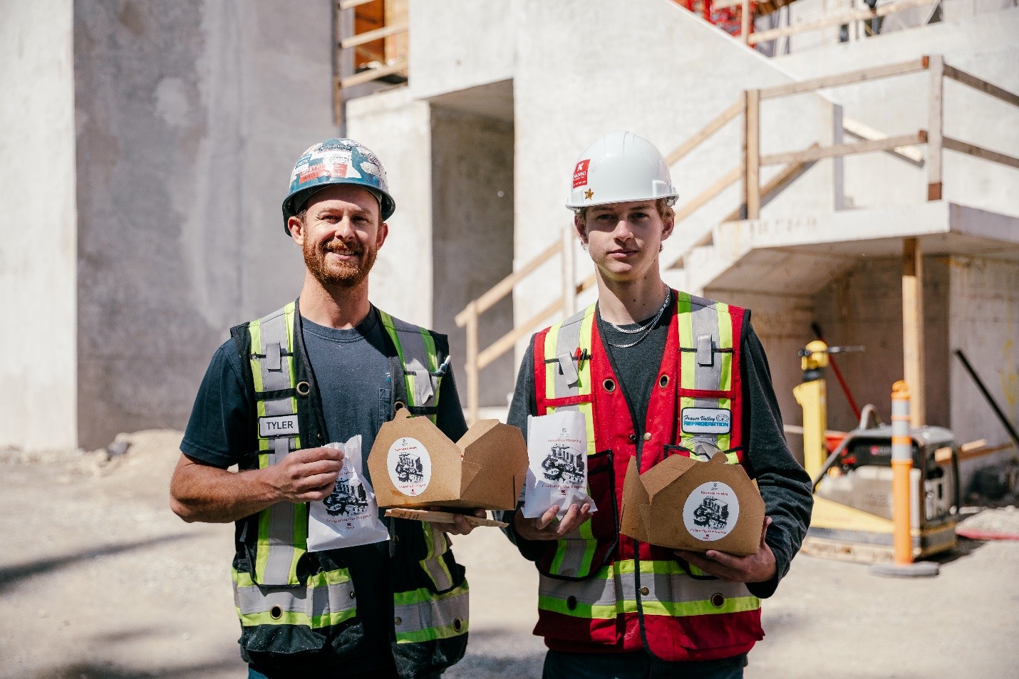 two guys wearing construction material standing in front of kwantlen hosuing holding boxes of indegenious food 